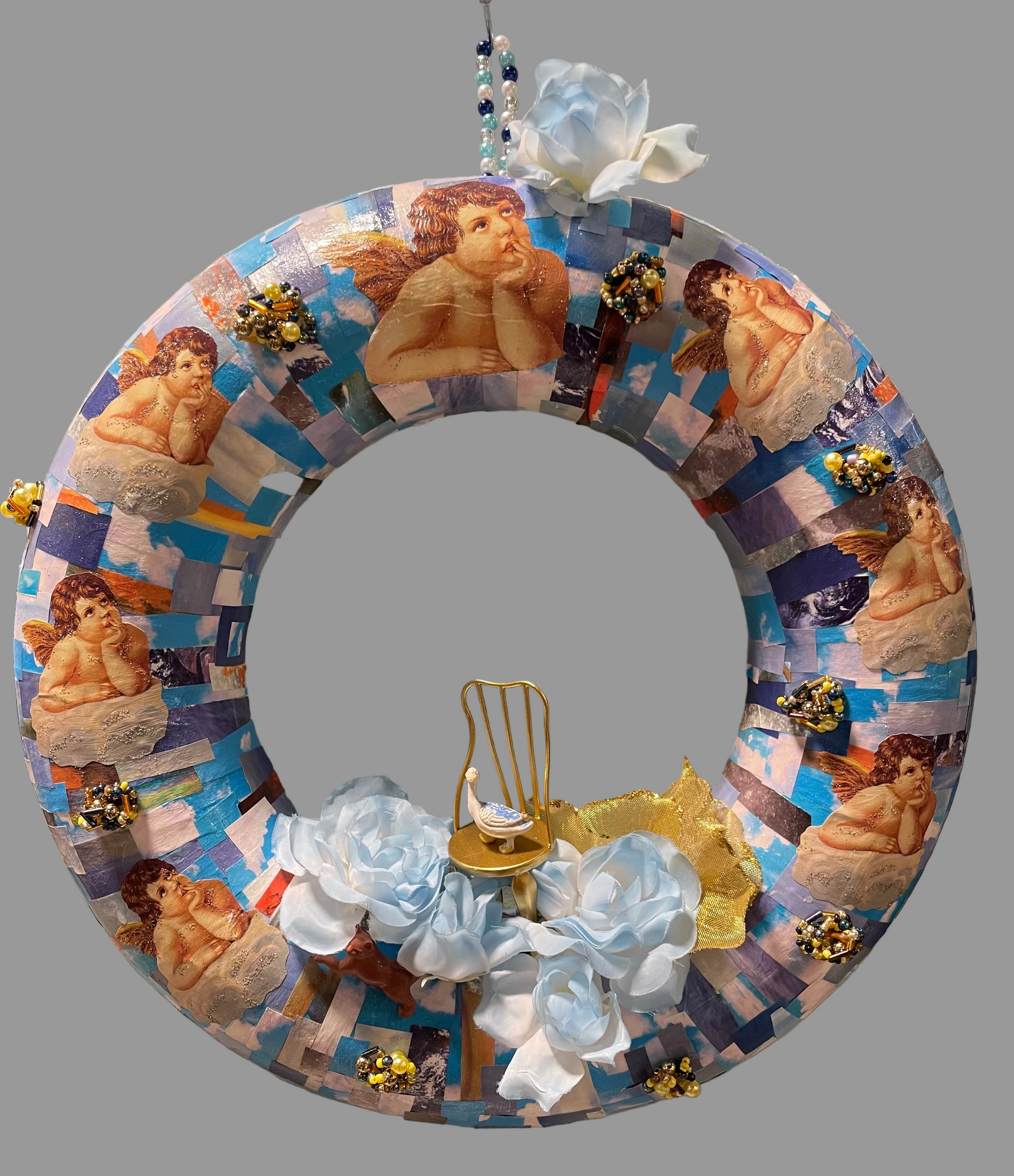A decorative circular wreath made from blue and white collage-style paper featuring classical female figures, embellished with light blue fabric flowers, a gold heart-shaped ornament, and small gold floral accents, photographed indoors against a light wall.